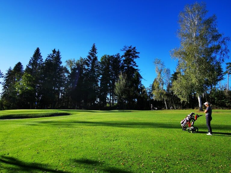 Eine Golfspielerin ist mit ihrem Golftrolley auf dem Golfplatz unterwegs.